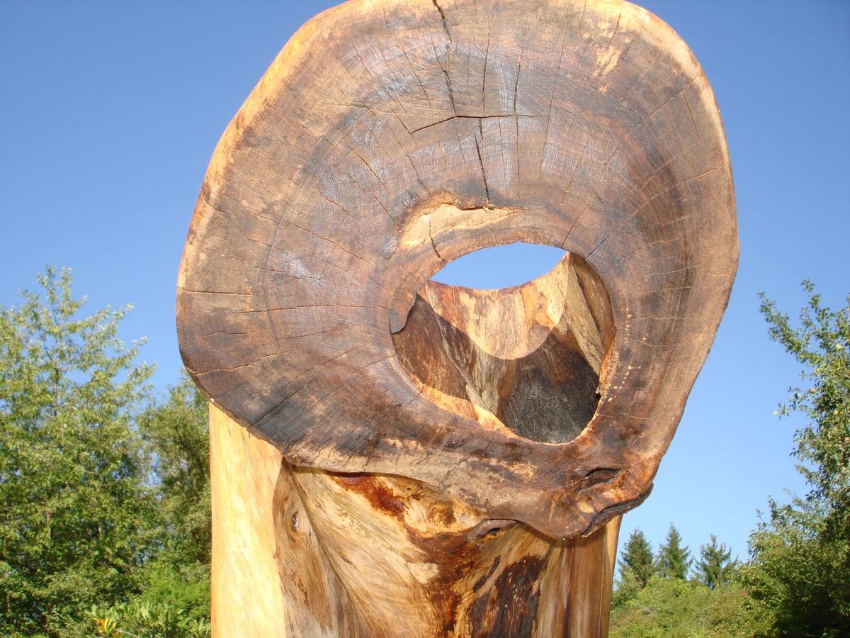 Eine hohe Holzplastik aus einem Baumstamm vor blauem Himmel und grünen Bäumen. Das obere Ende des Stammes ist als große, flache Scheibe ausgearbeitet, in deren Zentrum sich eine kreisförmige Öffnung befindet.