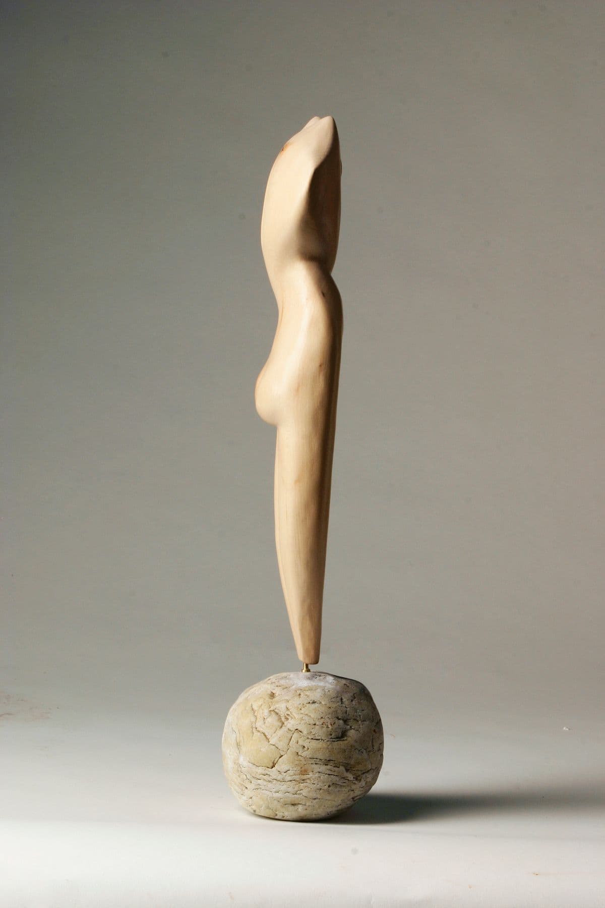 Eine schlanke, vertikale Plastik aus hellem, glatt poliertem Holz, die einen abstrahierten menschlichen Körper zeigt. Sie ist mit einem dünnen Metallstift auf einem runden, naturbelassenen Stein montiert; das Holz weist natürliche Merkmale wie kleine Astlöcher und einen feinen Trockenriss auf.