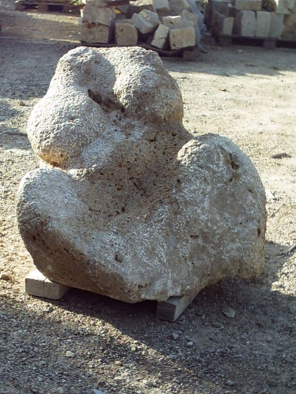 Eine massige, liegende Steinplastik aus grobporigem Tuffstein mit runden, organischen Ausformungen. Das Objekt ruht auf zwei Holzbalken auf einem Kiesboden in einem Außenlager.